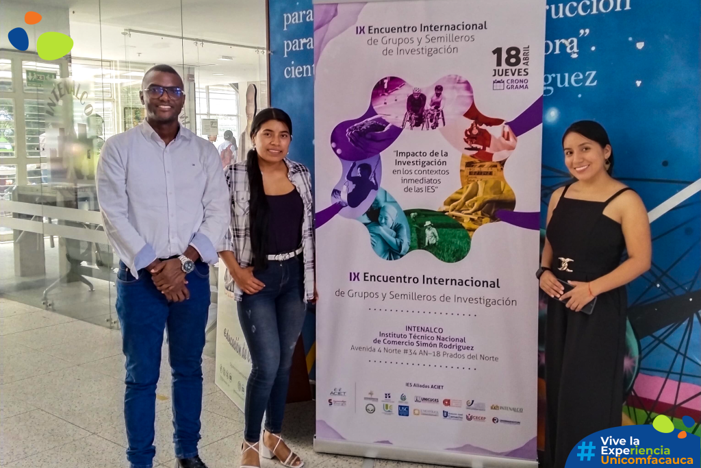 Dos estudiantes investigadoras y el profesor Diego Díaz posando frente a la publicidad del evento: IX Encuentro Internacional de Grupos y Semilleros de Investigación.