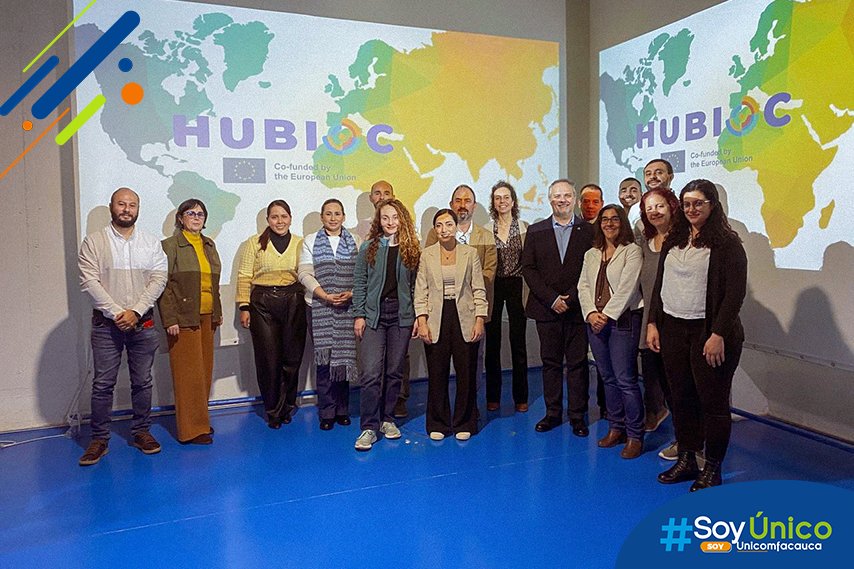 Destacada marca de agua Representantes de instituciones de Colombia, Argentina, España y Rumania, sonriendo a la cámara mientras se proyecta en la pared el logo de HUBIOC con un croquis del mapa mundi.