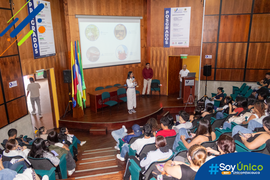 Foto general desde la parte de arriba del auditorio que lo muestra completamente lleno en lo que una de las invitadas imparte información en la tarima.