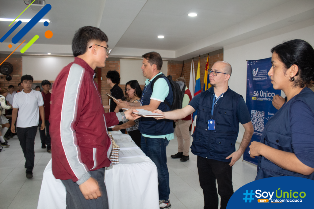 Estudiante participante de las olimpiadas, recibiendo por parte de un docente, un cuaderno personalizado del concurso.