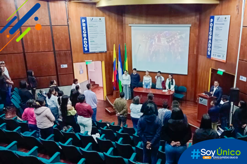 Fotografía de espaldas general del Auditorio Julio Arboleda, en donde los asistentes están de pie mirando hacia el frente en donde está ubicada la mesa principal conformada por: la Gestora Social de Popayán, Yessenia Velasco, nuestra Vicerrectora de Investigación y Extensión, Mg. Andrea Carolina Navas Calixto; el decano de nuestra Facultad de Ciencias Empresariales, Mg. Denis Augusto Lara Papamija; la Coordinadora del CEPRIDE, Ph.D. Juliana Liloy Valencia, y el director de nuestro programa de Contaduría Pública, Mg. Walter Ruano López.